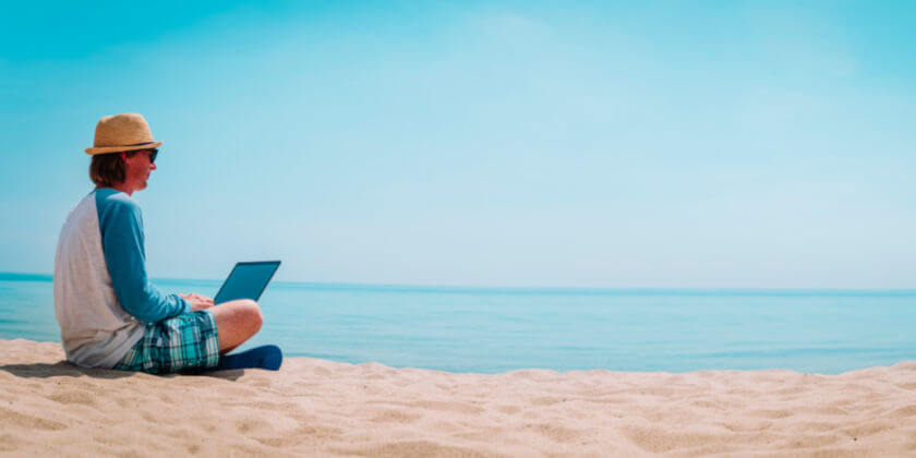A man with a laptop sits on the beach by the sea